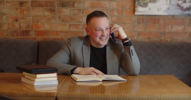 Ένας άντρας έχει μια χαρούμενη συζήτηση στο τηλέφωνο ενώ κάθεται σε ένα καφέ για ένα βιβλίο. — Αρχείο Βίντεο