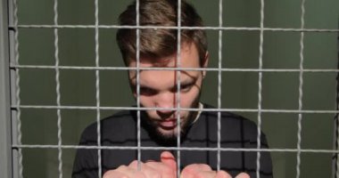 A man in handcuffs in the prison cell stands upset, holding on to the bars.