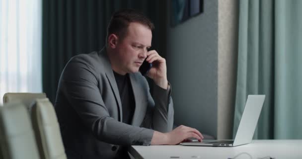 Σοβαρός άνθρωπος που εργάζεται σε φορητό υπολογιστή και μιλάει στο κινητό τηλέφωνο — Αρχείο Βίντεο