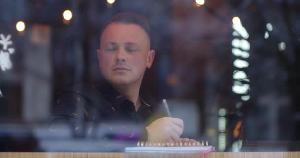 Seorang pria duduk di sebuah kafe yang nyaman, berpikir tentang teks dan menulisnya dalam sebuah buku catatan — Stok Video