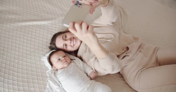 Mamãe se deita na cama com o bebê e tira uma selfie no telefone — Vídeo de Stock