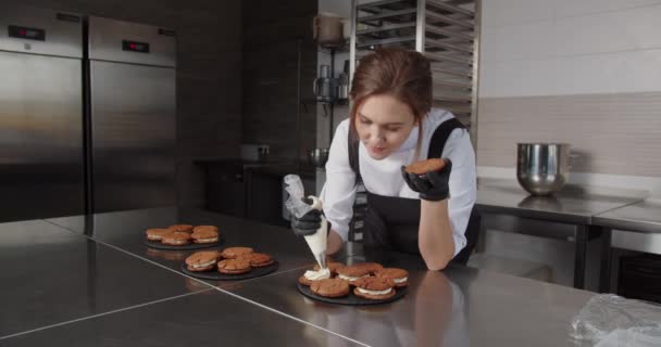 Com a ajuda de uma manga de confeitaria, uma garota de confeitaria aplica creme aos biscoitos. — Vídeo de Stock