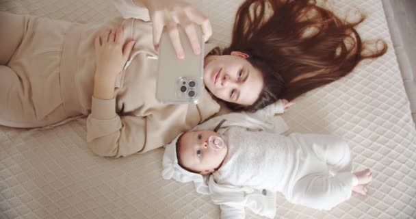 Young mother lies on the bed with the baby and takes a selfie on the phone — Stock Video