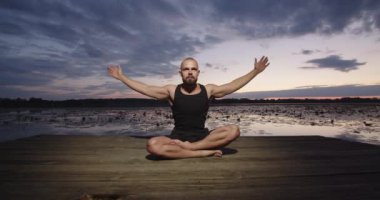 A man does yoga on the river bank during sunset in summer.
