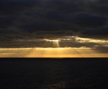 Denizin üzerinde gün doğumu, bulutlu gökyüzü ve bulutların arasında güneş ışığı.
