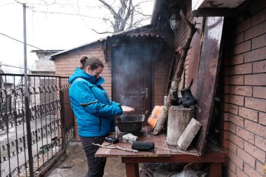 Mariupol, Ukrayna - 5 Mart 2022: Kadın Ukrayna 'nın Mariupol şehrinin bombardımanı altında evinin dışındaki şenlik ateşinin etrafında ısınıyor. Pencereler tahtalarla güçlendirilmiş. Telsiz masada..