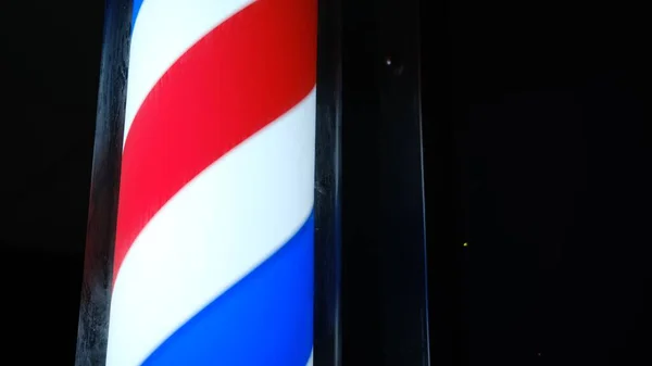 An old-styled barber shop sign, symbol. Round pole light in dark. Old-fashioned rotating led red white blue stripes pole light near men hair salon. Advertise of a small business outdoors. Hairdresser