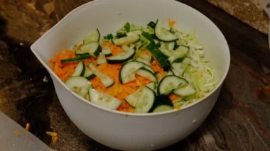 Old woman hands cook adding fresh cucumber slices in bowl with salad.