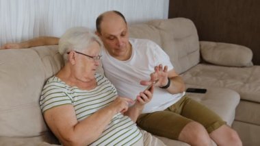 In living room on one side of sofa sitting old elderly 80s grandmother with adult 40s son, woman holding smartphone showing to older man new app, teach how use device help with usage concept.
