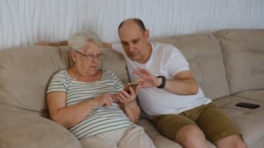 In living room on one side of sofa sitting old elderly 80s grandmother with adult 40s son, woman holding smartphone showing to older man new app, teach how use device help with usage concept.
