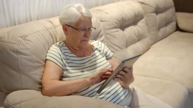 Pleasant senior older lady resting on couch, using applications on tablet. Happy old mature woman chatting in messenger or social network with friends or children, shopping in internet store.