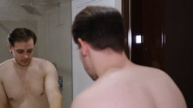 Banyoda sabah prosedürleri. Beyaz önlüklü genç bir adam aynanın karşısında duruyor. Derisi sorunlu ve bakımlı. Standart olmayan görünüm