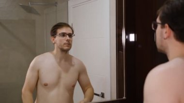Young man in glasses examining his face while watching into the mirror.