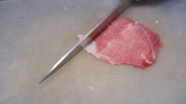 An old woman chops raw pork on a chopping board with a Knife.