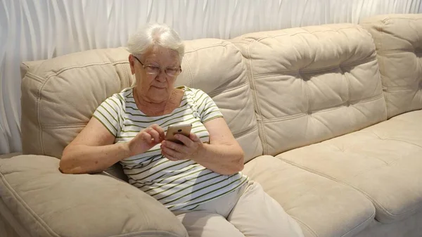 Pleasant senior older lady resting on couch, using applications on smartphone. Happy old mature woman chatting in messenger or social network with friends or children, shopping in internet store
