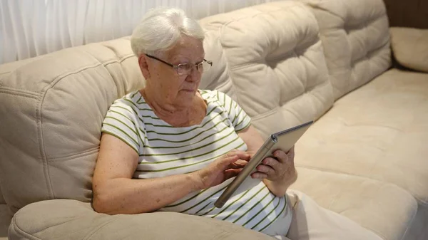 Pleasant senior older lady resting on couch, using applications on tablet. Happy old mature woman chatting in messenger or social network with friends or children, shopping in internet store.