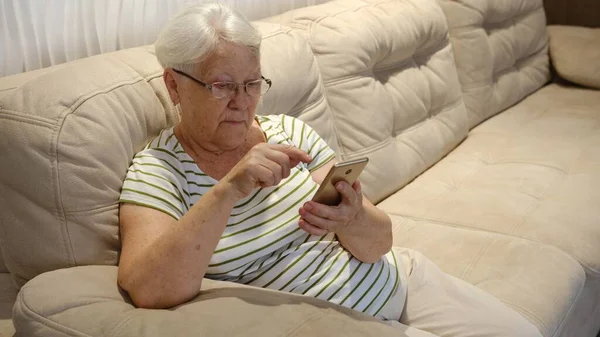 Pleasant senior older lady resting on couch, using applications on smartphone. Happy old mature woman chatting in messenger or social network with friends or children, shopping in internet store
