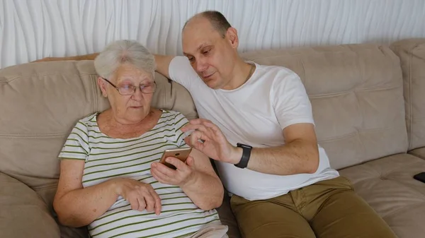 In living room on one side of sofa sitting old elderly 80s grandmother with adult 40s son, woman holding smartphone showing to older man new app, teach how use device help with usage concept.