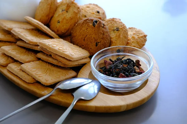Dried tea leaves with tea pot and cookies on a gray table with spoons and wooden board.