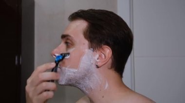 Serious young guy shaving his beard in a bathroom by razer.