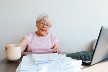 Mature editor reading printed text, making notes, using laptop, working from home, drinking coffee tea,