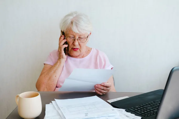 Senior woman holding papers busy at laptop managing house utility bills or finances, aged female using computer working with bank loan or mortgage documents online. Elderly and technology concept. talking to advisor by phone, working with documents a