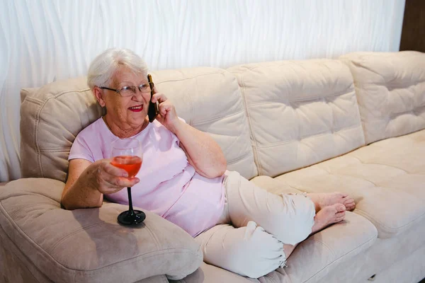 Photo of aged woman happy positive smile call talk cellphone drink alcohol wine rest relax weekend home. Senior woman at sofa talking by phone with glass of wine smiling