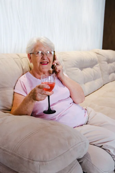 Photo of aged woman happy positive smile call talk cellphone drink alcohol wine rest relax weekend home. Senior woman at sofa talking by phone with glass of wine smiling