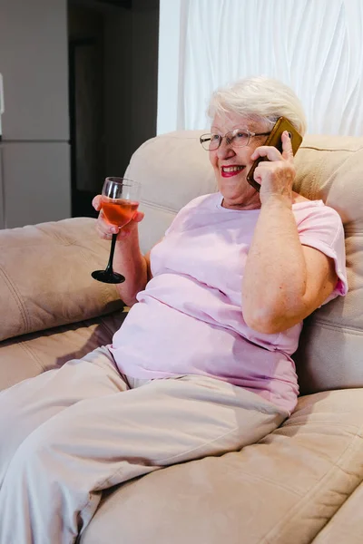 Photo of aged woman happy positive smile call talk cellphone drink alcohol wine rest relax weekend home. Senior woman at sofa talking by phone with glass of wine smiling