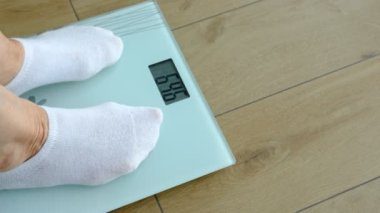 old woman standing on weight scale in living room, senior woman cheking her weight on digital scales.