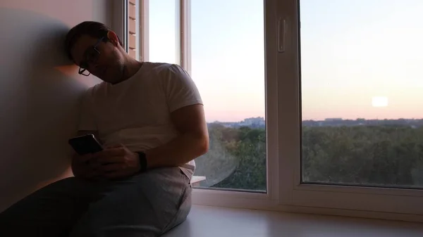 A silhouette of man sits by the window with a telephone and chating on the sunset light.