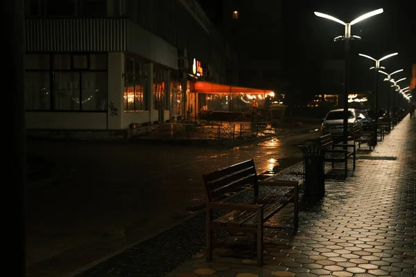 Rainy late evening in the park, a row of wooden benches, neon reflections of street lamps in wet asphalt.