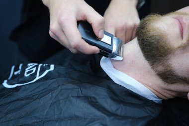 Profesyonel usta berber, müşterinin sakalını elektrikli makasla kesiyor. Berber dükkanında bir adamın sakalının kesimi. Berber Adamlar. Reklamcılık ve berber dükkanı konsepti.