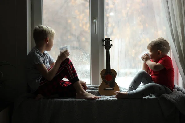 Çocuklar pencere eşiğinde oturuyorlar. Bardaktan çay iç ve pencereden dışarı bak. Beklenti, çocuk güvenliği ve plastik pencereler kavramı.