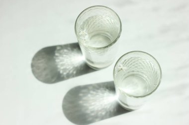 Temiz su ve keskin gölgeli iki bardak beyaz ahşap bir masada duruyor. Beyaz masa üzerinde su olan cam manzaralı.