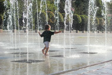 Islak elbiseli küçük bir çocuk parkta çeşmede koşuyor.