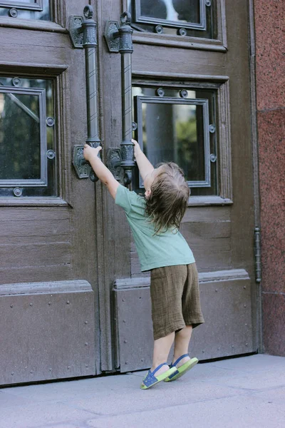 The boy stands with his back to the wooden door and pulls the handle. summer sunny day