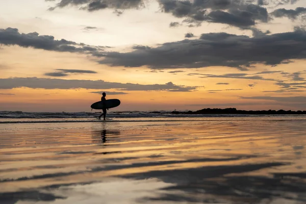 Sörfçü gün batımında sahilde sörf tahtasıyla yürüyor. Altın saatinde güzel renkler.