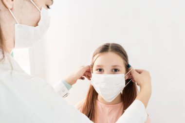 Bir hemşirenin ya da doktorun elleri çocuğa tıbbi maske taktı. Okula geri döndü. Yeni normal Coperid-19 durumu. Bir okul aç. Doktorda. Tıbbi maskeli Avrupalı kız çocuğu. Korunmak için tıbbi maske takan çocuklar.