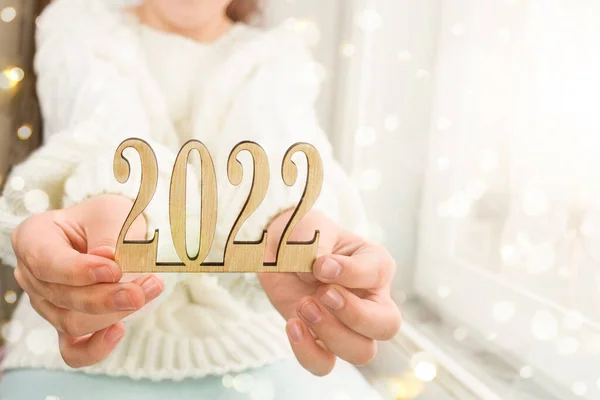 Beyaz bir kazakla pencerenin kenarında oturan ve 2022 yılının ahşap numaralarını tutan bir çocuğun elleri. Işığı söndürüyorum. Kişiliğini kaybetme. Seçici odaklanma. Tahta sayılar. Yeni yıla hazırlanıyorduk. Yeni yılı kutluyoruz. 