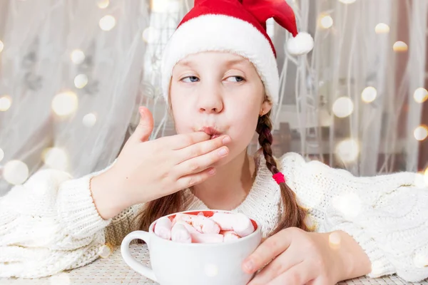 Noel Baba şapkalı bir kız çocuğu masaya oturur ve ellerinin parmaklarını yalayarak bir kupa sıcak çikolatadan marşmelov yer. Seçici odaklanma. Odaklanamayan ışık. Noel içkileri ve tatlılar. Saha Sahasının Derinliği Samimi Odaklanma Anı