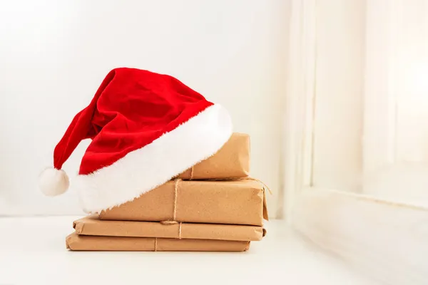 Noel Baba şapkalı hediye kutuları beyaz arka planda, pencerenin yanında duruyor. Seçici odaklanma. Noel ve Yeni Yıl için hediyeler hazırlama. Ekolojik Paketleme.