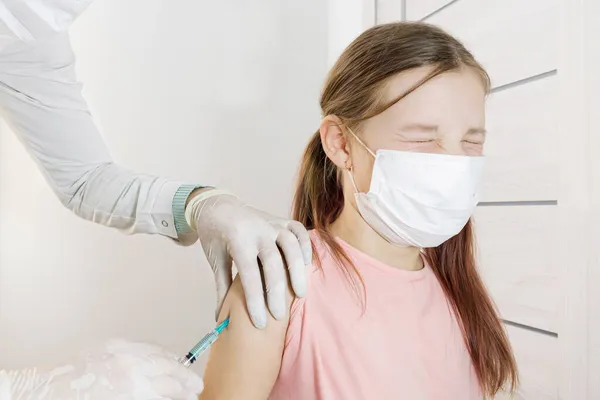 Tıbbi eldivenli eller, aşı sırasında gözlerini korkudan kapayan bir kızın omzuna enjeksiyon yaptı. Seçici odaklanma. Çocukluk aşıları. Doktorun hastanedeki randevusunda iğneler ve prosedürler. 