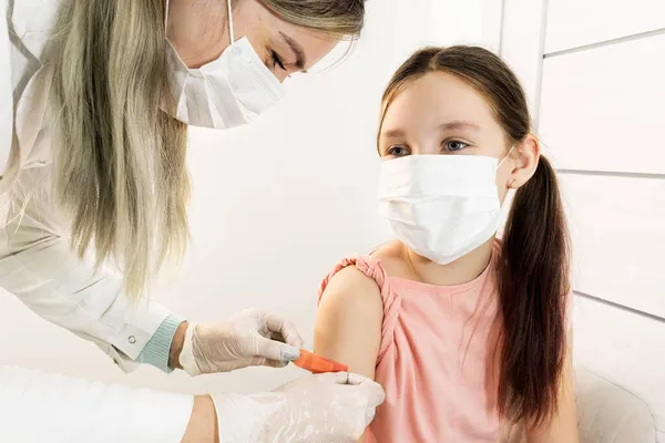 Lastik eldivenli ve koruyucu maskeli bir hemşire, çocukların aşılanması ve enjeksiyonlar için yakınlarda oturan bir çocuğun omzuna tıbbi sıvayı yapıştırıyor. Hastanedeki Doktor 'un resepsiyonundaki prosedürler..