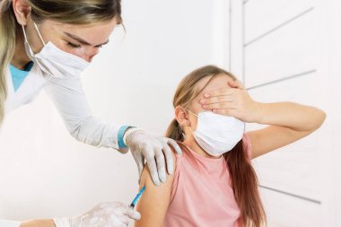 Tıbbi maskeli ve üniformalı bir hemşire korkusundan gözlerini kapatan bir çocuğun omzunda aşı yapıyor. Seçici odaklanma. Çocukların aşılanması. Çocuk aşıları ve enjeksiyonlar doktorun ofisinde..