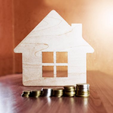 Ahşap ev sembolü, ön planda bozuk para yığınları bulunan ahşap bir yüzey üzerinde durmaktadır. Kamu hizmetlerinin ödemesi, ahşap sembol evi, ev satın alma ve satış konsepti. İpotekler ve ev kredileri. Aile ve barınma.