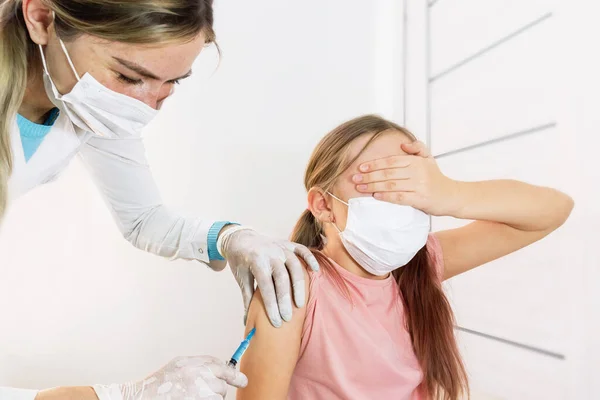 Tıbbi maskeli ve üniformalı bir hemşire korkusundan gözlerini kapatan bir çocuğun omzunda aşı yapıyor. Seçici odaklanma. Çocukların aşılanması. Çocuk aşıları ve enjeksiyonlar doktorun ofisinde..