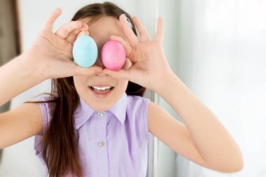 Gözlerinin yanında 2 Paskalya yumurtası tutan ve gülümseyen kız çocuğu. Seçici odaklanma. Uzayı kopyala Mutlu Paskalyalar. Çocuklar ve Paskalya yumurtaları