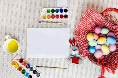 Boyalı Paskalya yumurtaları ve oyuncak bir tavşan pencerenin yanındaki ahşap bir sepetin üzerinde beyaz bir çarşafın yanında uzanıyorlar. Top View Mocup. Seçici odak ve beyaz arkaplan.