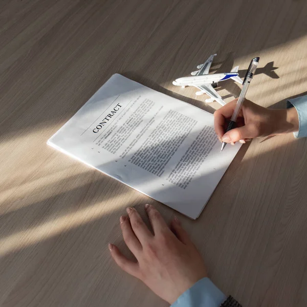 Uçuş sigortası, seyahat sigortası. sözleşmeli uçak. Uçak ve hava ekipmanları kiralanıyor. Yakın plan. Kadın eli bir kalem tutuyor ve imzalamadan önce sözleşmeyi inceliyor, belgenin yanında uçağın sembolü yatıyor ve güneş ışığı parlıyor. 